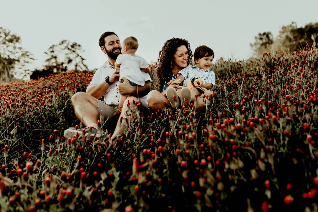 Family of four with a baby girl and toddler boy sitting In a patch of red flowers playing with the kids. Pediatrician Macon GA