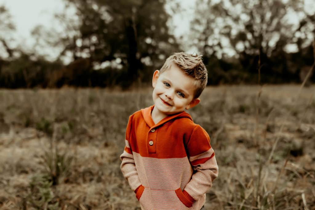 Family Photo Session Checklist: What to Do + What to Bring 4 Young boy in an orange and tan sweater smiling in a grassy field during a family photo session.