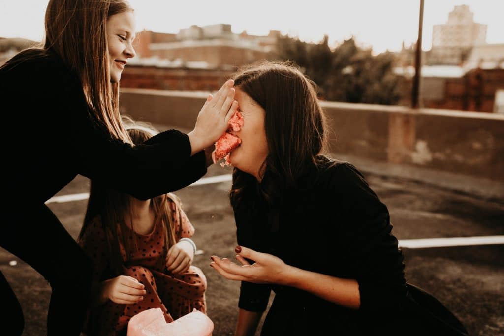 two daughters smashing pink cake into moms face on a city rooftop. 