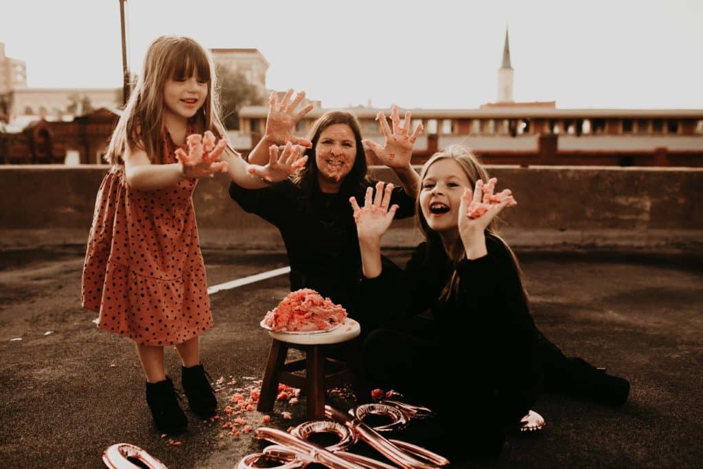mom and two daughters holding their hands up with cake smashed on their hands for a birthday session. 