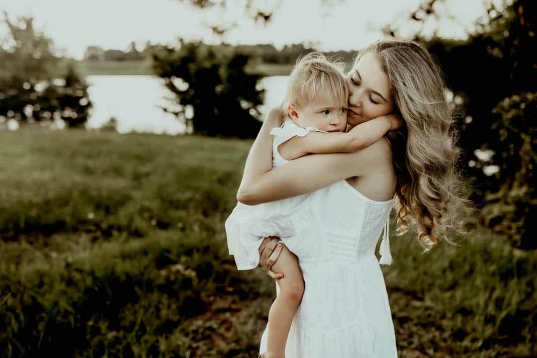 Mom with long blonde hair holding and hugging baby girl in her arms in a field with a lake background
