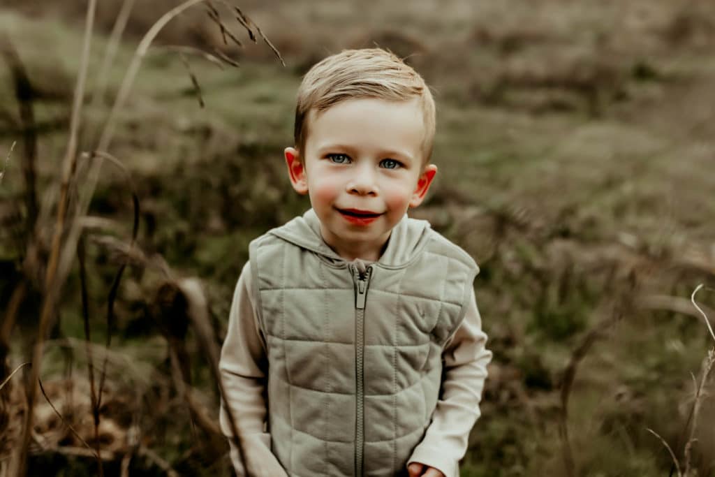 Toddler boy smiling at the camera while wearing a sage vest.