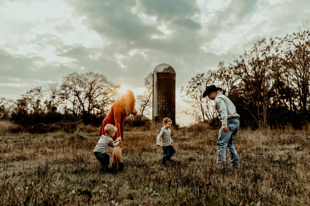 family of 4 chasing one another in a field at sunset
