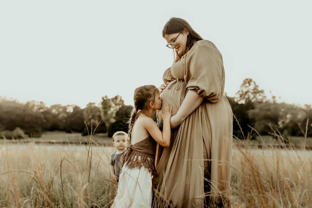 How I Photograph Maternity Sessions When You Don’t Feel “Glowy” 2 Pregnant mother standing in a field while her daughter kisses her baby bump, capturing a quiet moment of family connection during a maternity session in Middle Georgia.