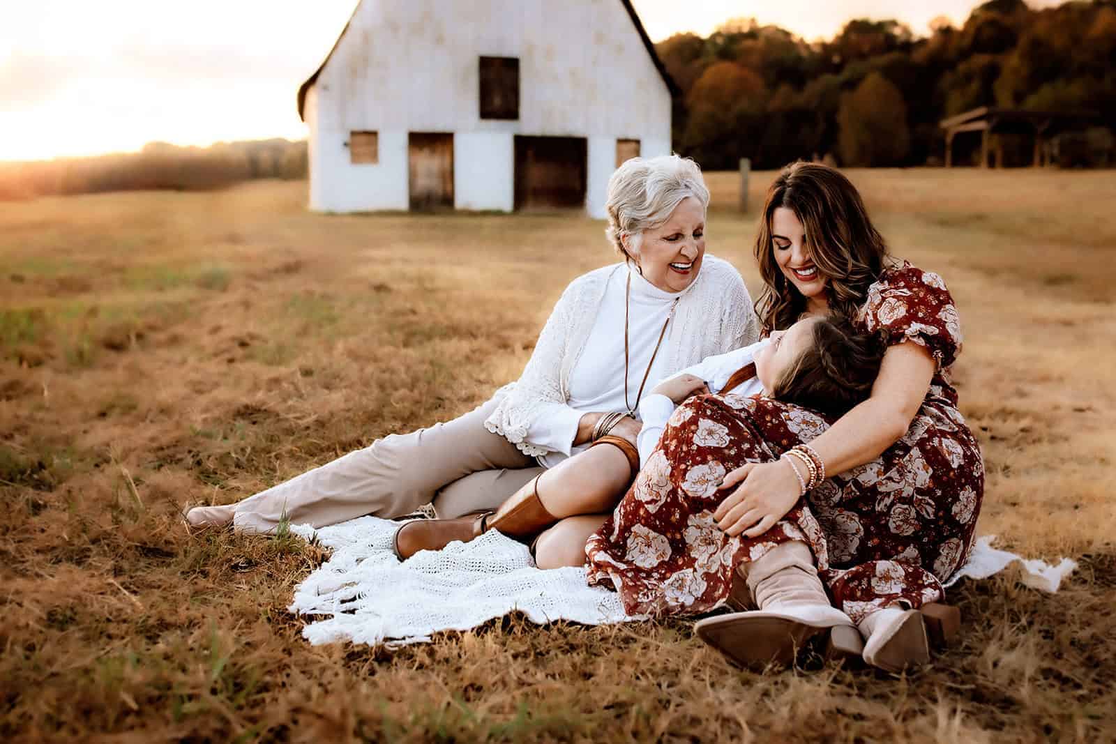 A Generational Motherhood Session at Vaughters Farm in Stonecrest, GA 1 Generational motherhood session at Vaughters Farm in Stonecrest, GA, with a mother, grandmother, and daughter sitting together in a field at sunset.