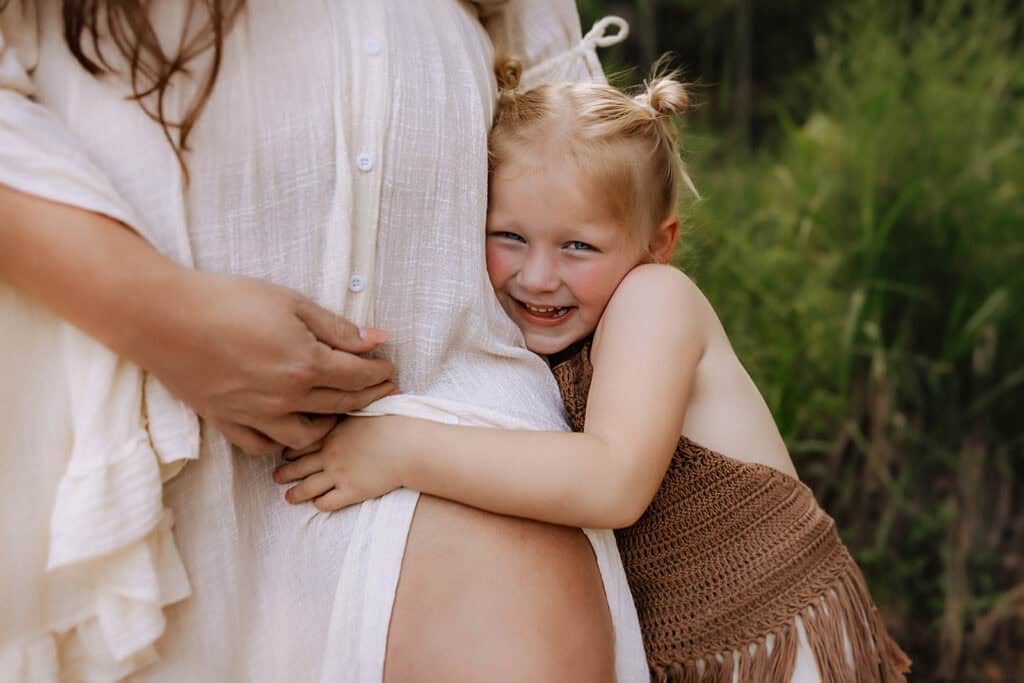 Best Time to Schedule Maternity Photos (So You Actually Feel Like Yourself) 4 Young child hugging their mother’s baby bump during an outdoor maternity photo session
