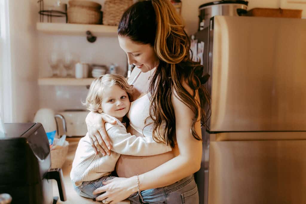 Best Time to Schedule Maternity Photos (So You Actually Feel Like Yourself) 14 Pregnant mother holding her toddler close during an in-home maternity photo session