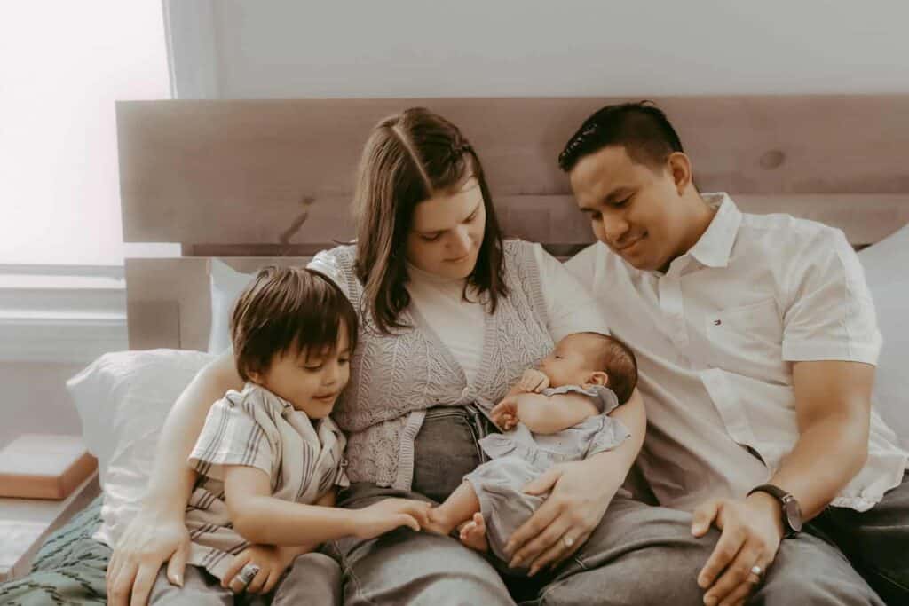 Do Newborn Photos Have to Happen in the First Two Weeks? 10 Parents and older brother sitting together on the bed with their newborn baby.