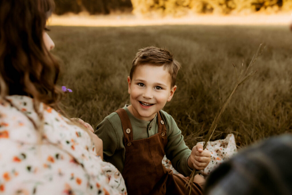 How Long Does a Family Photo Session Take? 3 Young boy smiling while sitting with his family in a grassy field during family photos